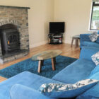 Living room with stone fireplace and log burning stove, blue rug in front with glass coffee table on the rug and two blue settees around the fireplace