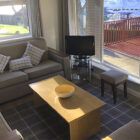 Fawn coloured setttees set around a coffee table with view through large window of decking and lake