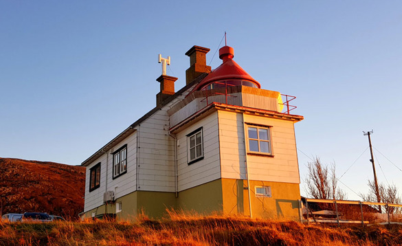 White lighthouse building with red lantern roof on a grassy hill at sunset