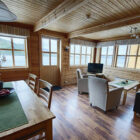 Living/dining room in a cabin with settee, two armchairs and table with dining chairs and view across a fjord in Norway