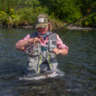 Lady stands waist-deep in a river, successfully landing a fish on a fly fishing line.