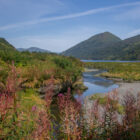 A winding river flows through a lush green valley toward a larger body of water, flanked by rolling green mountains under a pale blue sky.