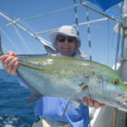 A smiling man wearing a sun hat and sunglasses on a boat, holding up a bright Bluefin Trevally with iridescent blue and green markings against a clear blue sky and ocean background.