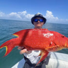 A man on a boat wearing a dark bucket hat and sunglasses, holding up a vibrant, bright red coral trout with blue spots and yellow-edged fins, set against a calm ocean and a blue sky with scattered clouds.
