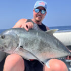 A smiling man in a light blue cap and sunglasses sitting on a boat and holding a large, silvery Giant Trevally across his lap, with the calm blue ocean and horizon in the background.