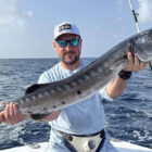A man on a boat holding a long, silvery fish with dark spots and sharp teeth against a blue ocean background.