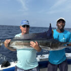 Gemini said Two men on a boat holding a large, silvery sailfish with its prominent dark blue dorsal fin extended, set against the backdrop of a vast, deep blue ocean