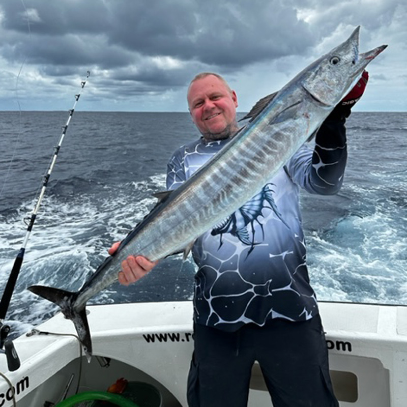 A man smiles while holding a large, slender wahoo fish on a boat