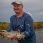 A man wearing a baseball cap and a blue long-sleeved fishing shirt smiling while holding a tiger fish he caught in Botswana. The fish features distinctive dark horizontal stripes and bright orange-red fins, with a colorful fishing lure still in its mouth, set against a background of a river and marshy shoreline under an overcast sky.
