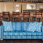 The interior of a lodge featuring a bright blue and white patterned sofa in the foreground and a long wooden bar in the background with intricate African carvings on its panels and high-backed wooden stools.