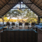 An open-air lounge at a river lodge featuring comfortable blue and patterned sofas, wooden coffee tables, and a row of chairs on a wooden deck overlooking a river and lush trees under a high thatched roof with a ceiling fan.