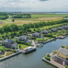 Aerial view of waterfront holiday homes along a canal with docked boats and green fields in the background.
