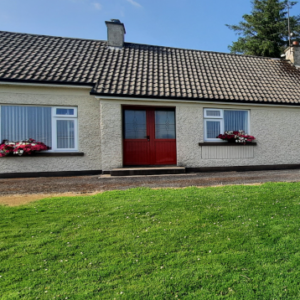 Front view of a cream-colored bungalow with a red door, dark tiled roof, and a large green lawn.