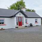 A white single-story cottage with a red front door and a dark grey tiled roof on a gravel driveway.
