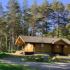 A wooden log cabin nestled in a dense pine forest under a clear blue sky.