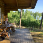An elderly man relaxing on the wooden porch of a log cabin overlooking a forest and lake.