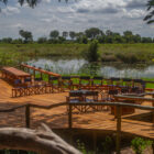 An expansive wooden viewing deck with safari chairs arranged around a central fire pit overlooking a marshy wetland.