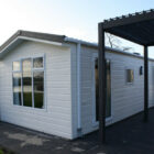 Exterior of a white mobile home chalet with dark window frames and a black pergola structure on a paved patio.