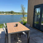 Outdoor patio dining set with a table and four chairs next to a modern wooden chalet overlooking a lake.
