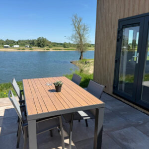 Outdoor patio dining set with a table and four chairs next to a modern wooden chalet overlooking a lake.