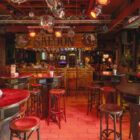 Rustic western-style saloon bar interior with high top tables, red stools, and hanging disco balls.