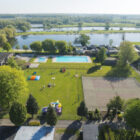 Aerial view of a holiday park featuring a swimming pool, tennis court, playground, and lakeside chalets.
