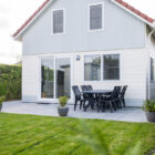 Modern white holiday chalet with sliding glass doors, a stone patio, outdoor dining table, and lawn.