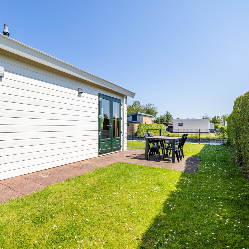Side exterior of a white wooden bungalow featuring double glass doors leading out to a small lawn and patio table.