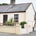 Front view of a cream-coloured cottage with window flower boxes, a wooden front railing, and a ramp leading up to the side entrance.