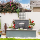 Modern outdoor water feature set against a white garden wall surrounded by potted flowers and climbing greenery.